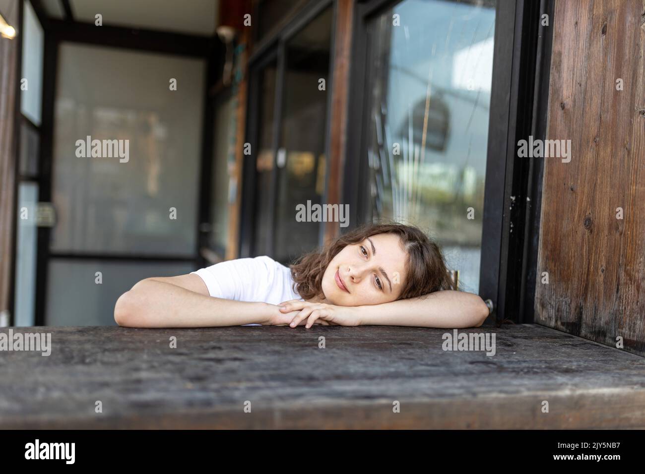 A smiling brunette girl in a white T-shirt, blue jeans sits at a wooden counter in a cafe, resting her chin in her hands. Vertical frame. Stock Photo