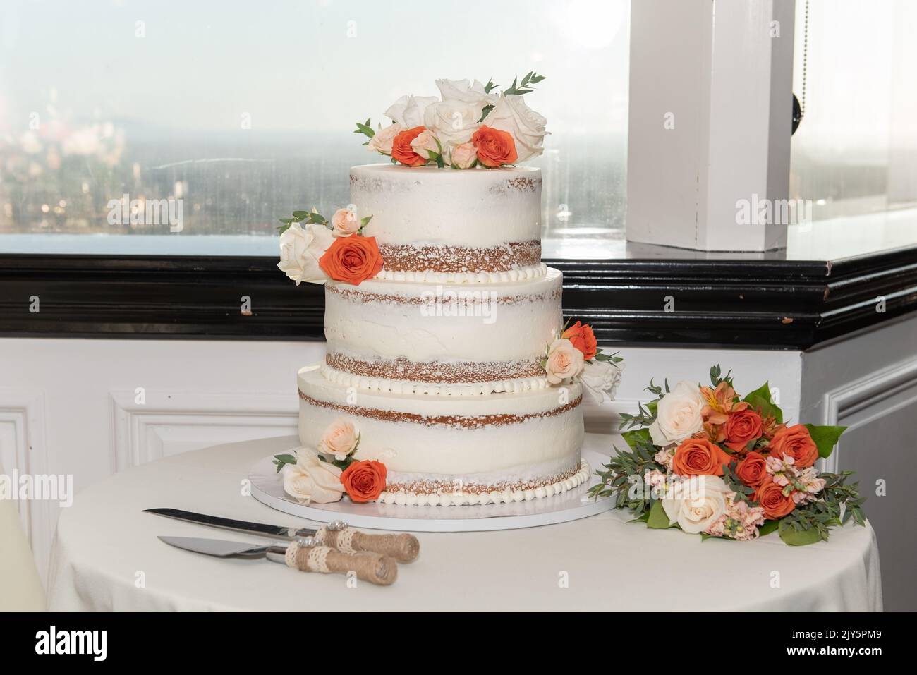 Triple layer wedding cake is set next to city view window along with serving set and bouquet of flowers. Stock Photo