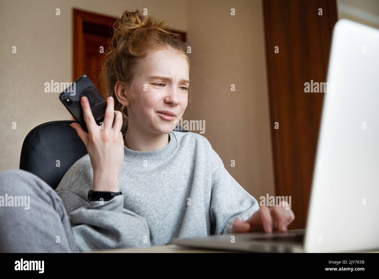 Caucasian red-haired beautiful teenage girl in a grey hoodie and pants, sitting at a table in front of a laptop with a smartphone. A girl with funny Stock Photo