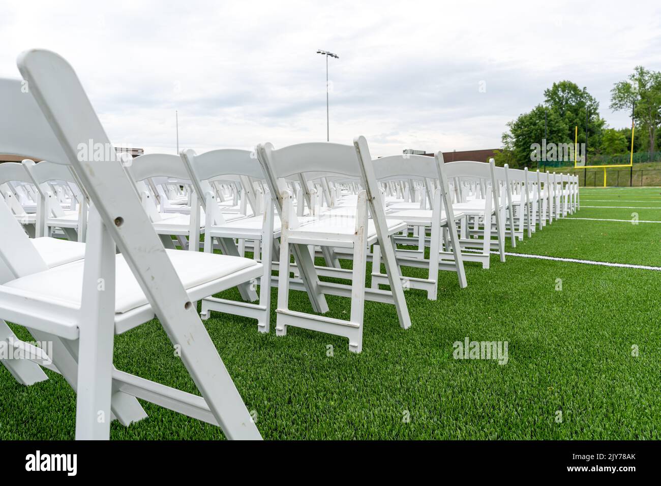 White chairs set-up in rows on a green synthetic turf athletic field for a high school graduation ceremony. Stock Photo