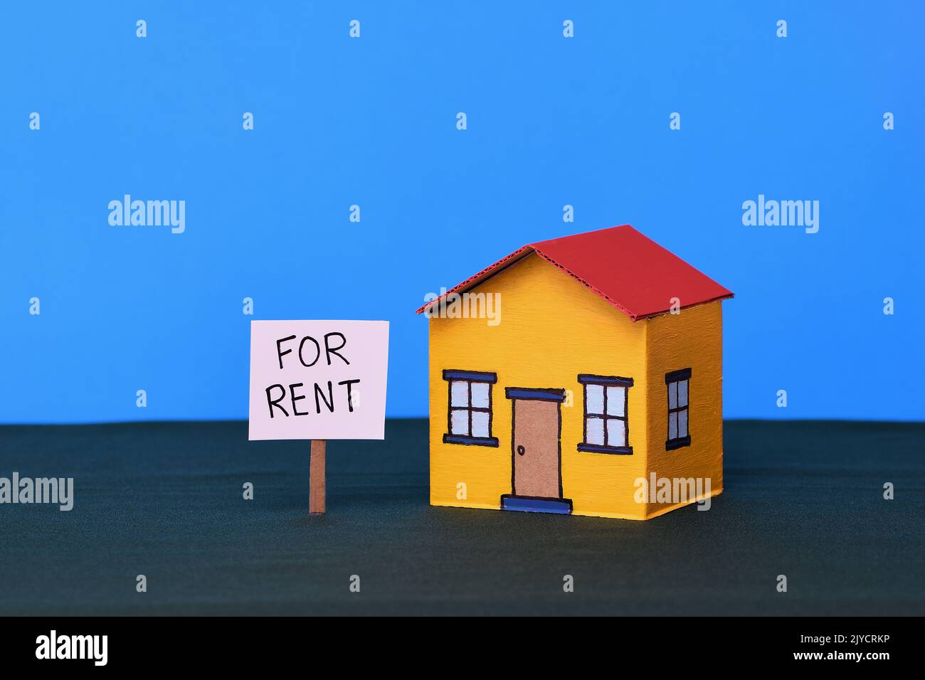 A yellow and red toy cardboard house in the middle of frame on a dark green base and blue background in soft lighting with a Realestate For Rent sign Stock Photo