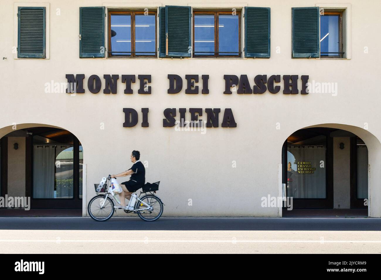 A woman cycling in front of the Monte dei Paschi di Siena Italian bank, Castiglione della Pescaia, Grosseto, Tuscany, Italy Stock Photo