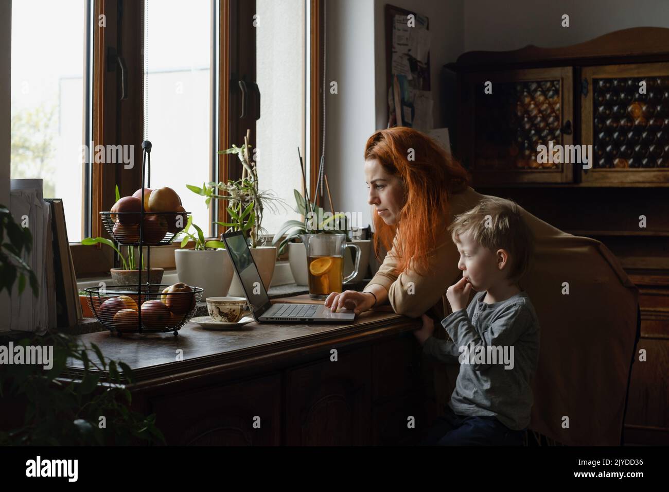 Ginger haired woman on kitchen with child looking in laptop. Vintage house, cozy family life. Boy with mother at home. Family morning Stock Photo