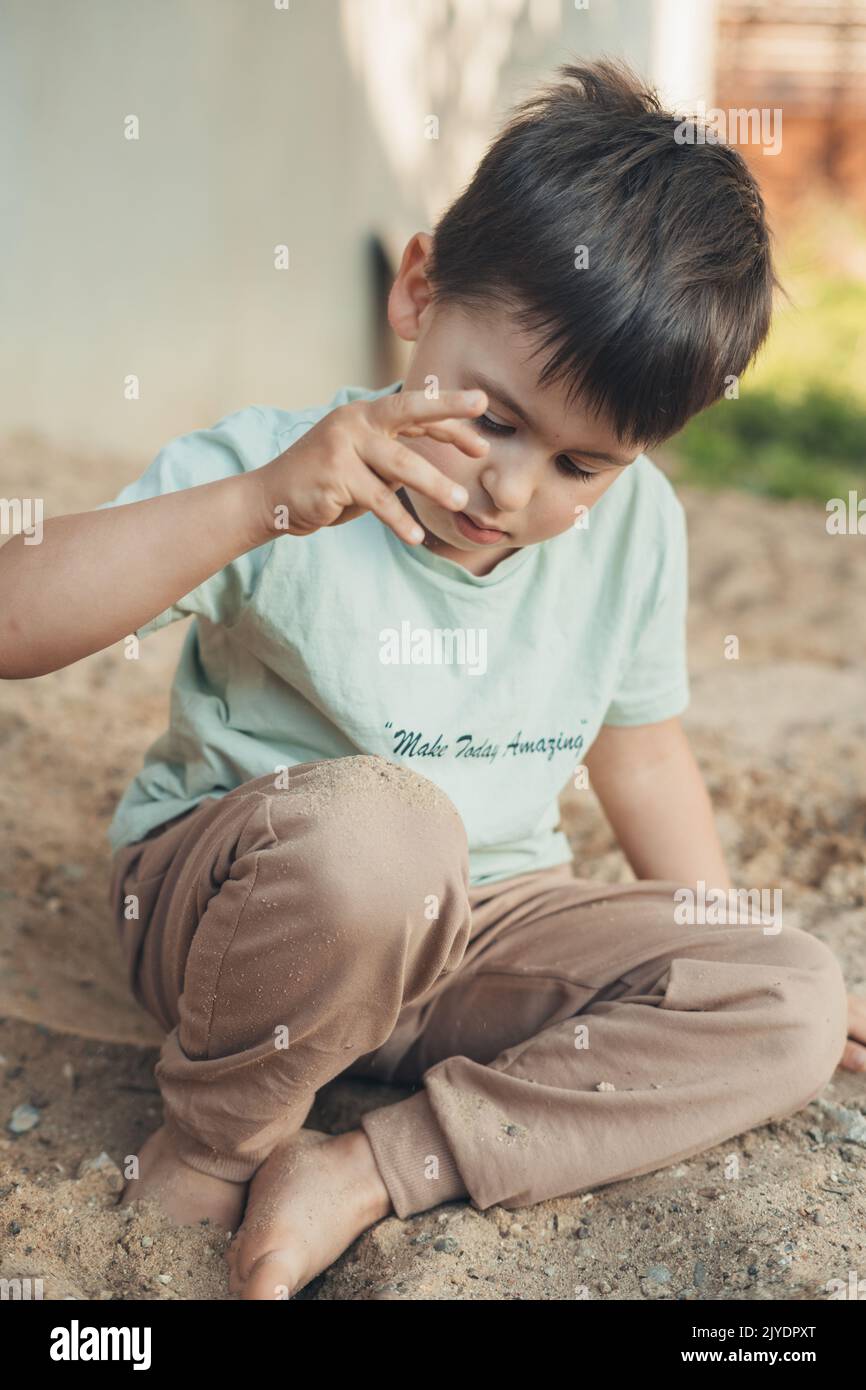 Boy carefully examining the sand flowing from his fist while playing outside in the garden. Creative play for kids. Quarantine with small kid. Stock Photo