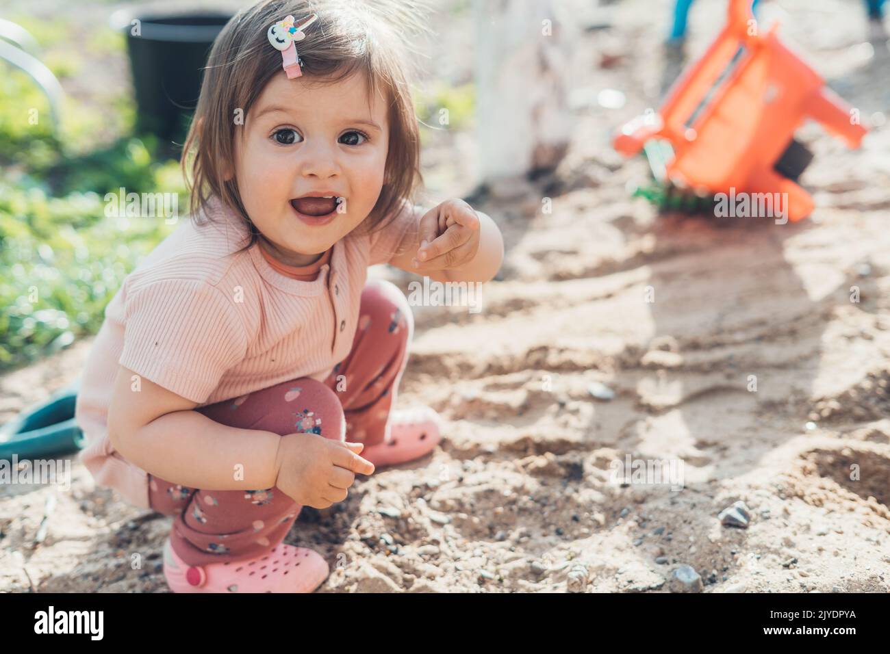 Adorable baby girl sitting on the playground and pointing to the camera what she discovered. Creative play for kids concept. Happy family, childhood Stock Photo