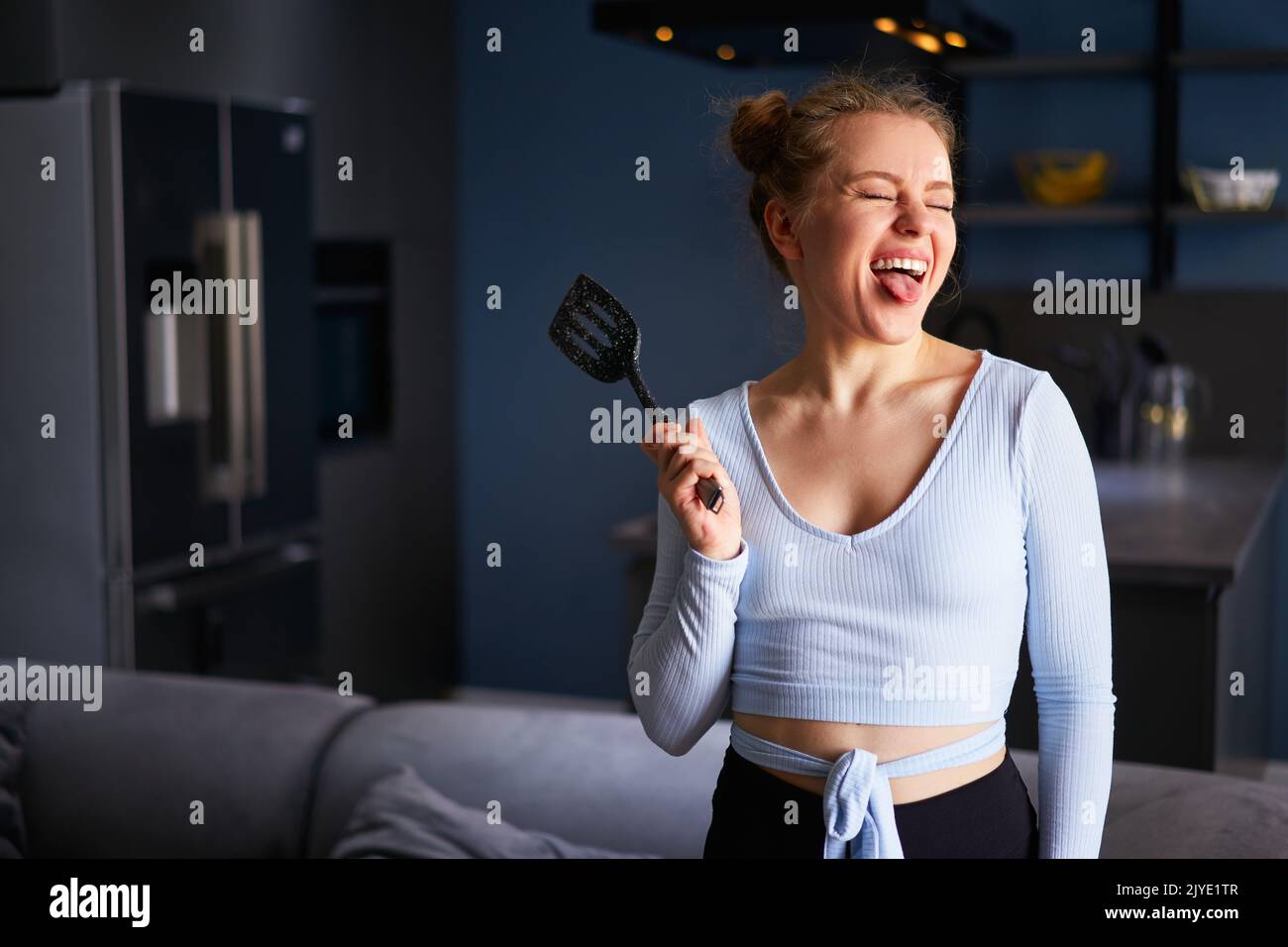 Young caucasian blonde woman holding a black spatula sticks out tongue with her eyes closed laughing with teeth standing at home on dark modern Stock Photo
