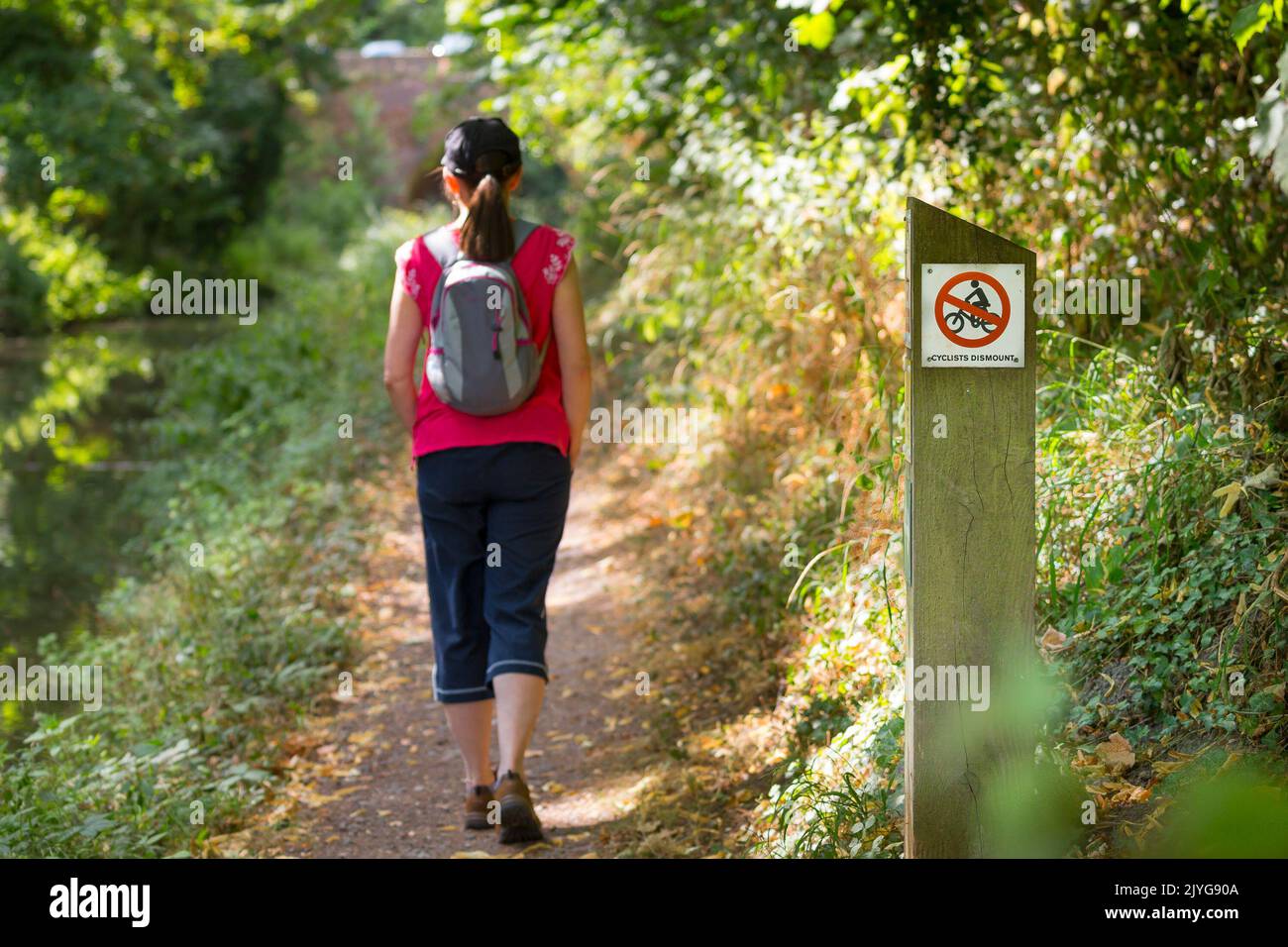 Rear view of a woman walking past a cyclist dismount sign on a canal towpath. Stock Photo
