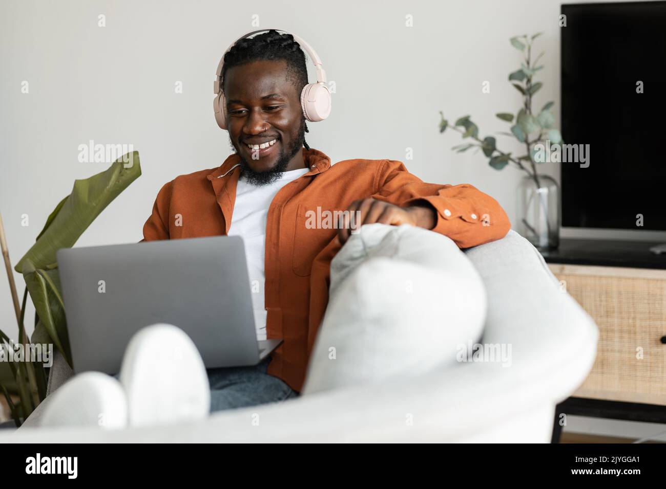 Positive black freelancer man in headphones working on laptop at home, sitting on couch in living room, free space Stock Photo