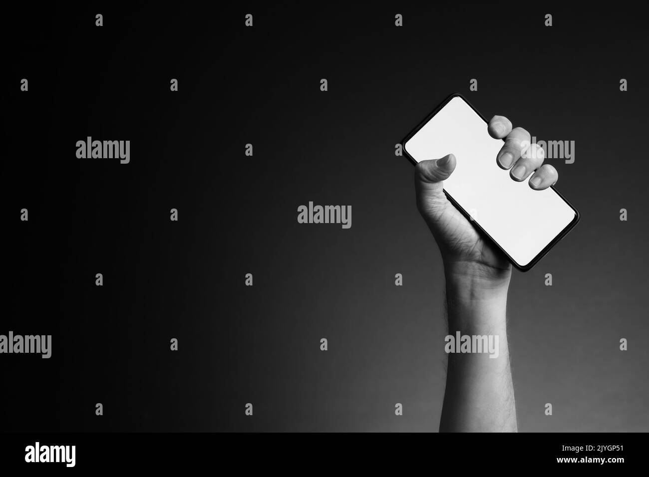 Black and white image of man's hand holding smart phone with blank white screen isolated on dark background with copy space Stock Photo