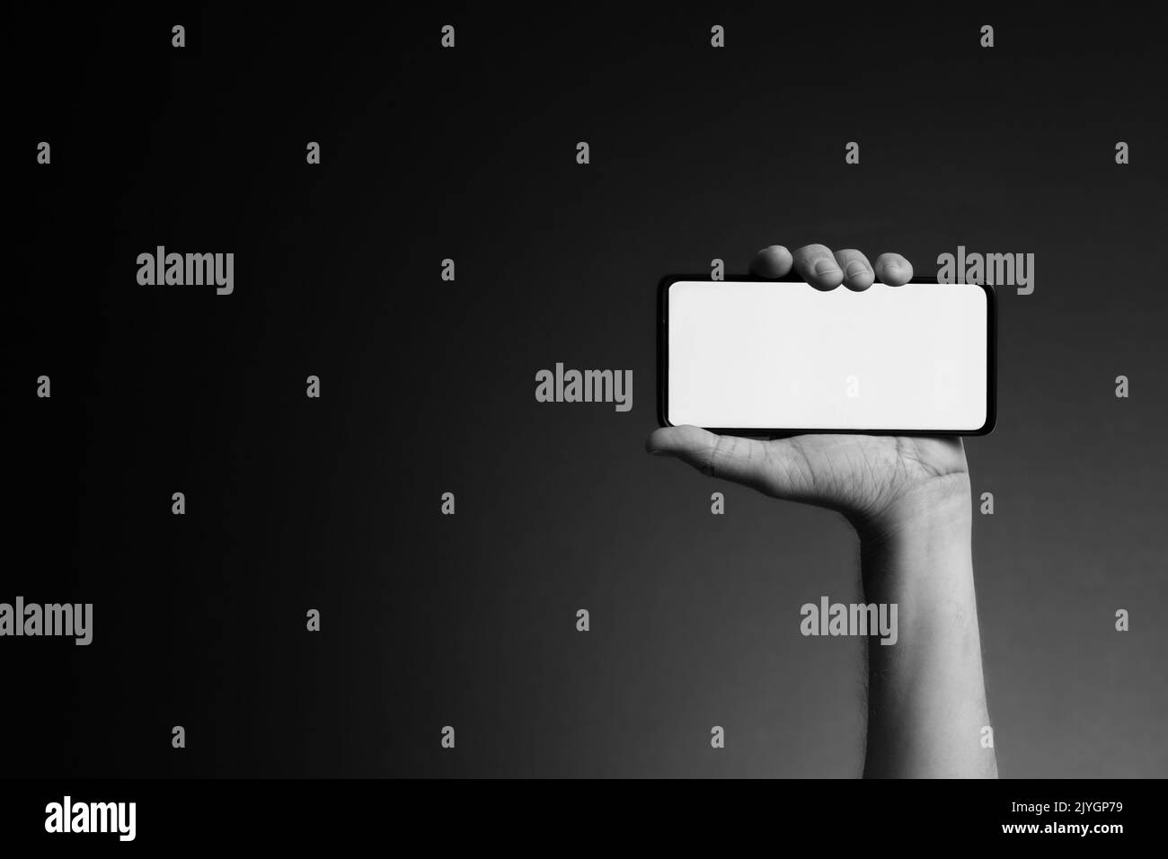 Black and white image of man's hand holding smart phone horizontally with blank white screen isolated on dark background with copy space Stock Photo