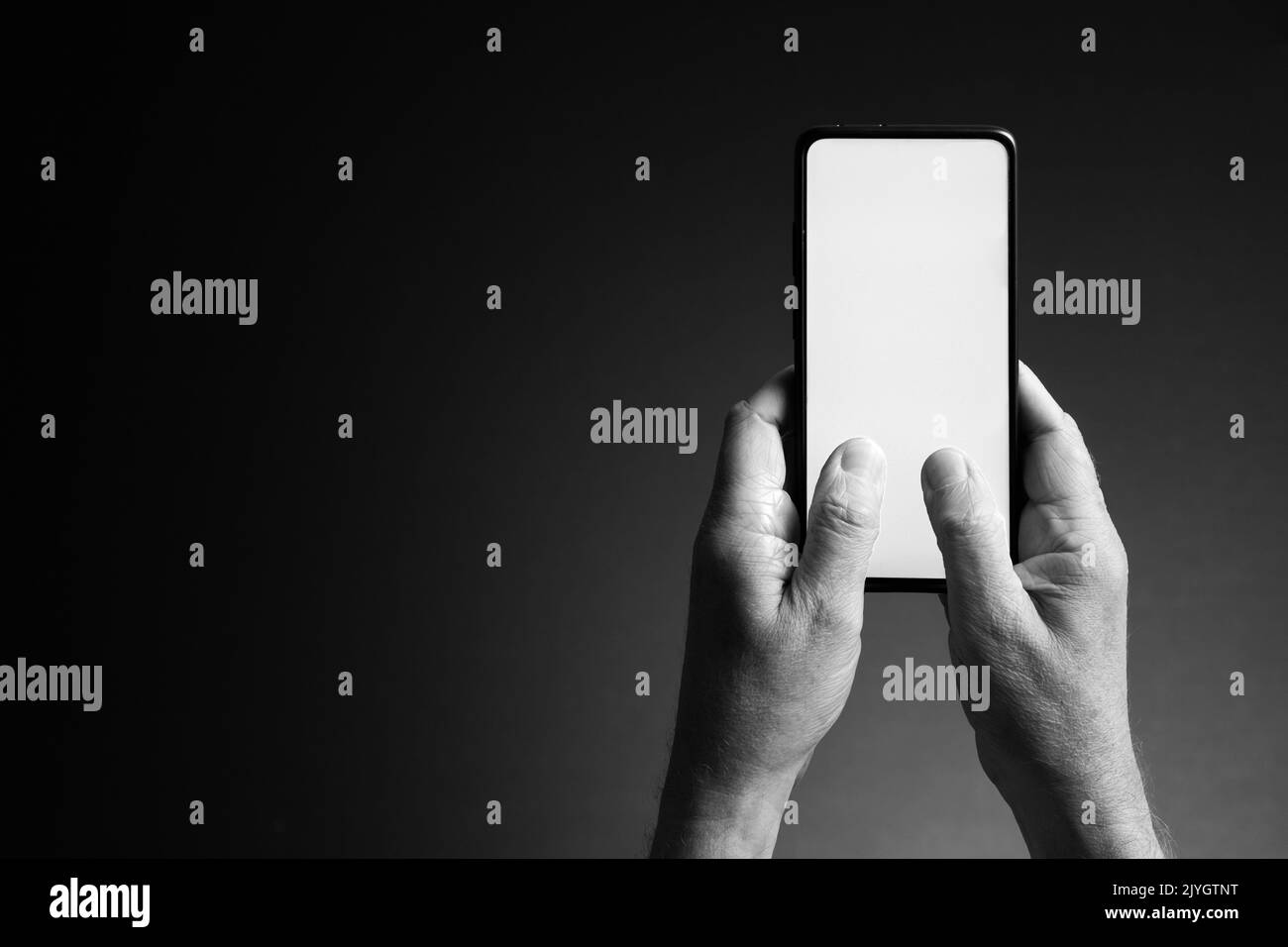 Black and white image of man's hands holding smart phone and texting with blank white screen isolated on dark background with copy space Stock Photo
