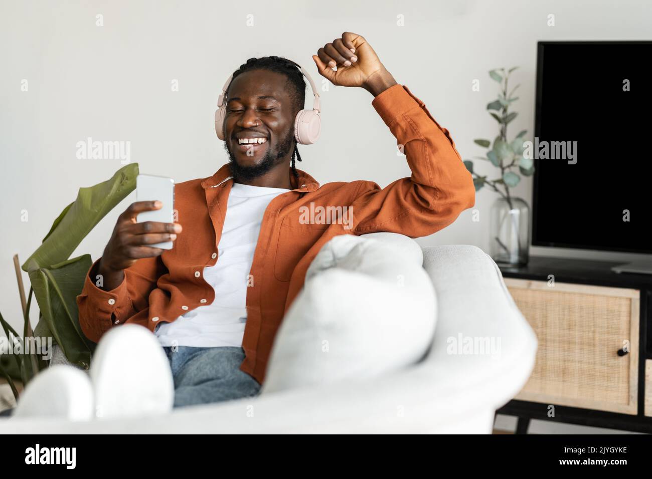 Yes. Overjoyed black man using smartphone and shaking clenched fist, celebrating victory, sitting on sofa Stock Photo