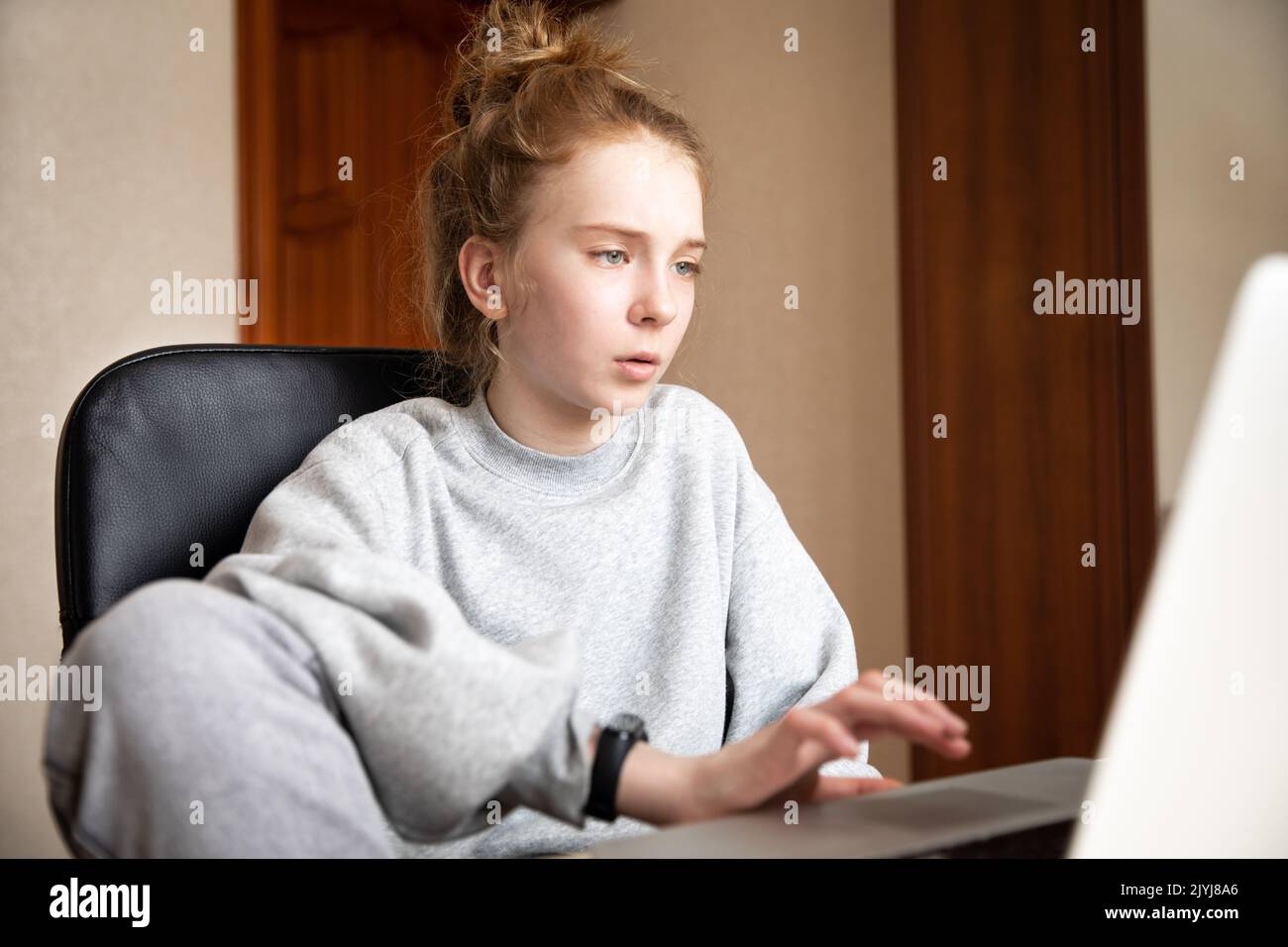 Caucasian cute red-haired teenage girl in a grey hoodie and pants, sitting at a table in front of a laptop indoors. Home distance education. Obtain Stock Photo