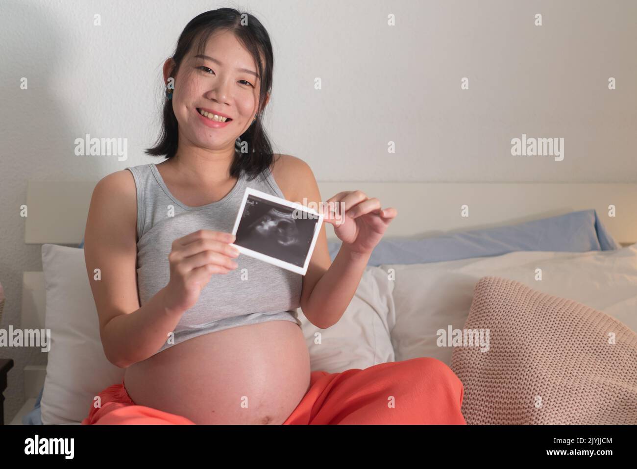 lifestyle home portrait of young happy and beautiful Asian Chinese woman pregnant sitting on bed holding ultrasound photo excited in pregnancy and mat Stock Photo