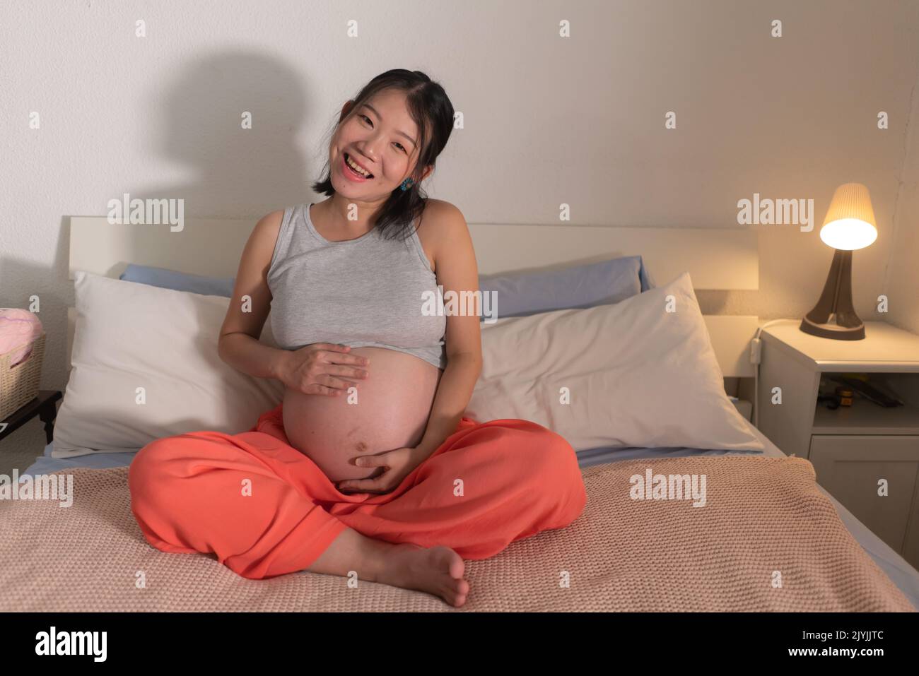 lifestyle home portrait of young happy and beautiful Asian Chinese woman pregnant sitting on bed relaxed and excited about maternity touching her bell Stock Photo
