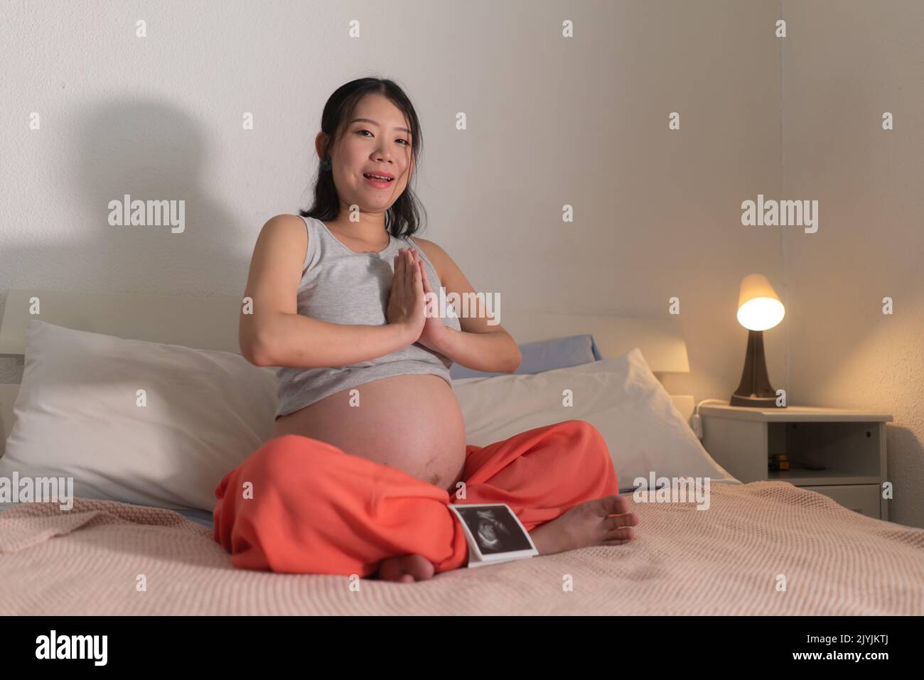 lifestyle home portrait of young happy and beautiful Asian Japanese woman pregnant doing relaxing yoga exercise on bed in maternity and pregnancy conc Stock Photo