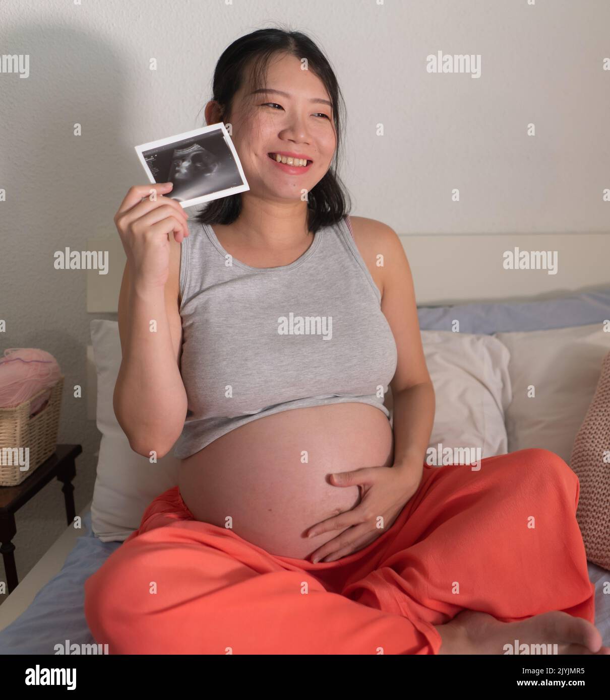 lifestyle home portrait of young happy and beautiful Asian Japanese woman pregnant sitting on bed holding ultrasound photo excited in pregnancy and ma Stock Photo