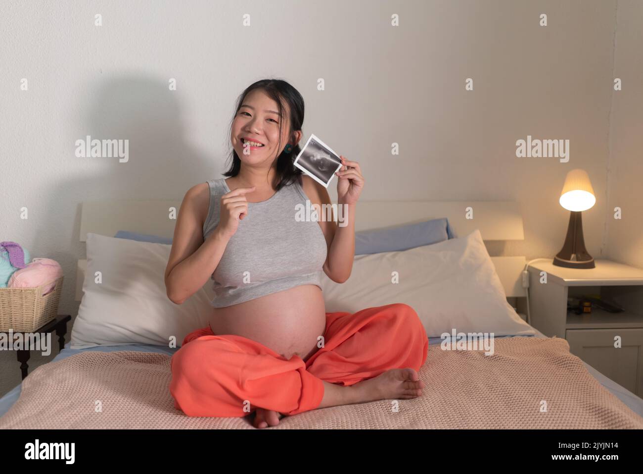 lifestyle home portrait of young happy and beautiful Asian Japanese woman pregnant sitting on bed holding ultrasound photo excited in pregnancy and ma Stock Photo