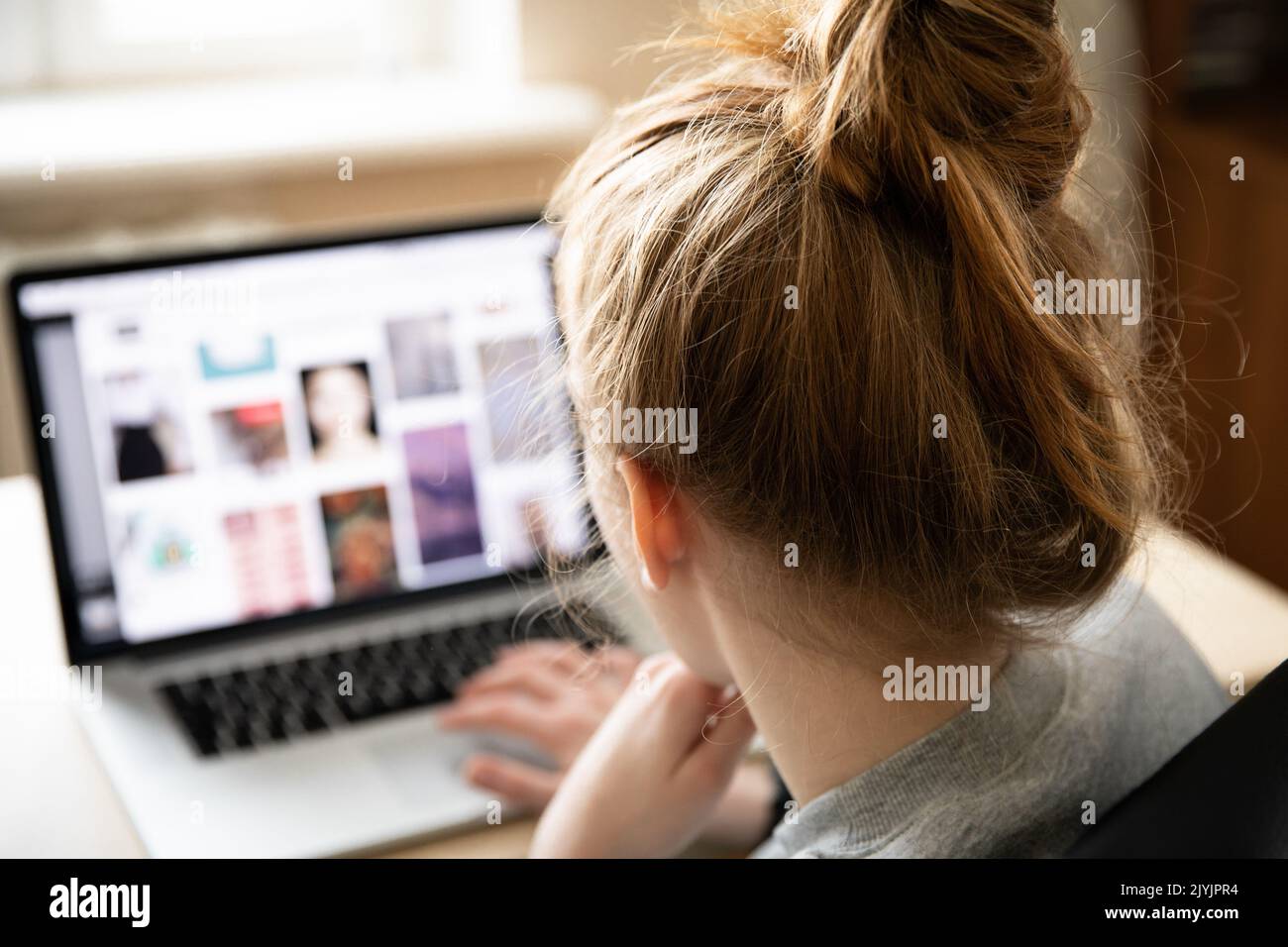 Red-haired girl in a grey hoodie sits at a light-colored table in front of a laptop against a window. A girl in a relaxed position looks at the Stock Photo