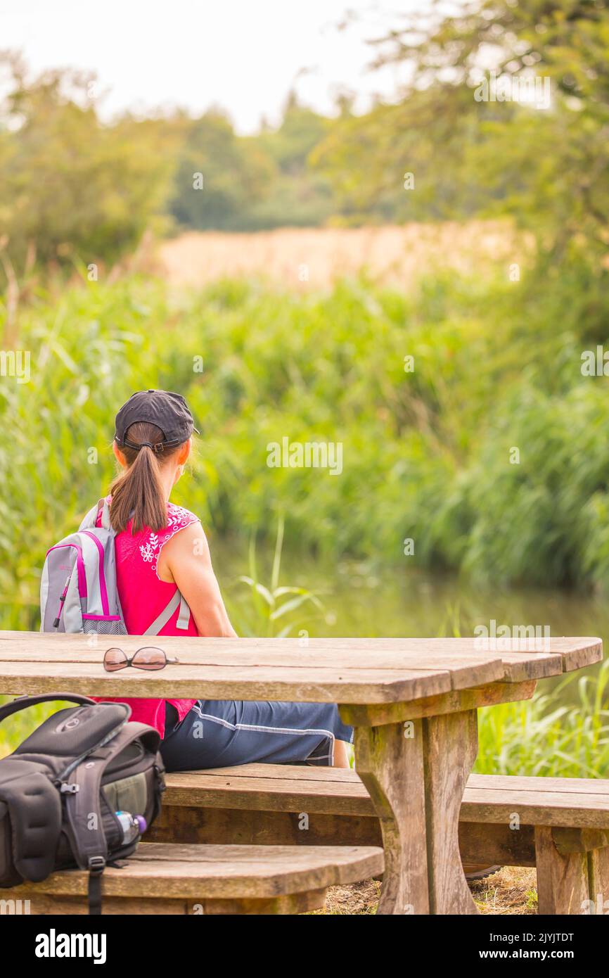 Rear view of young woman with ponytail wearing sports cap, sitting on a picnic bench in the countryside looking at a UK canal during warm  summertime. Stock Photo
