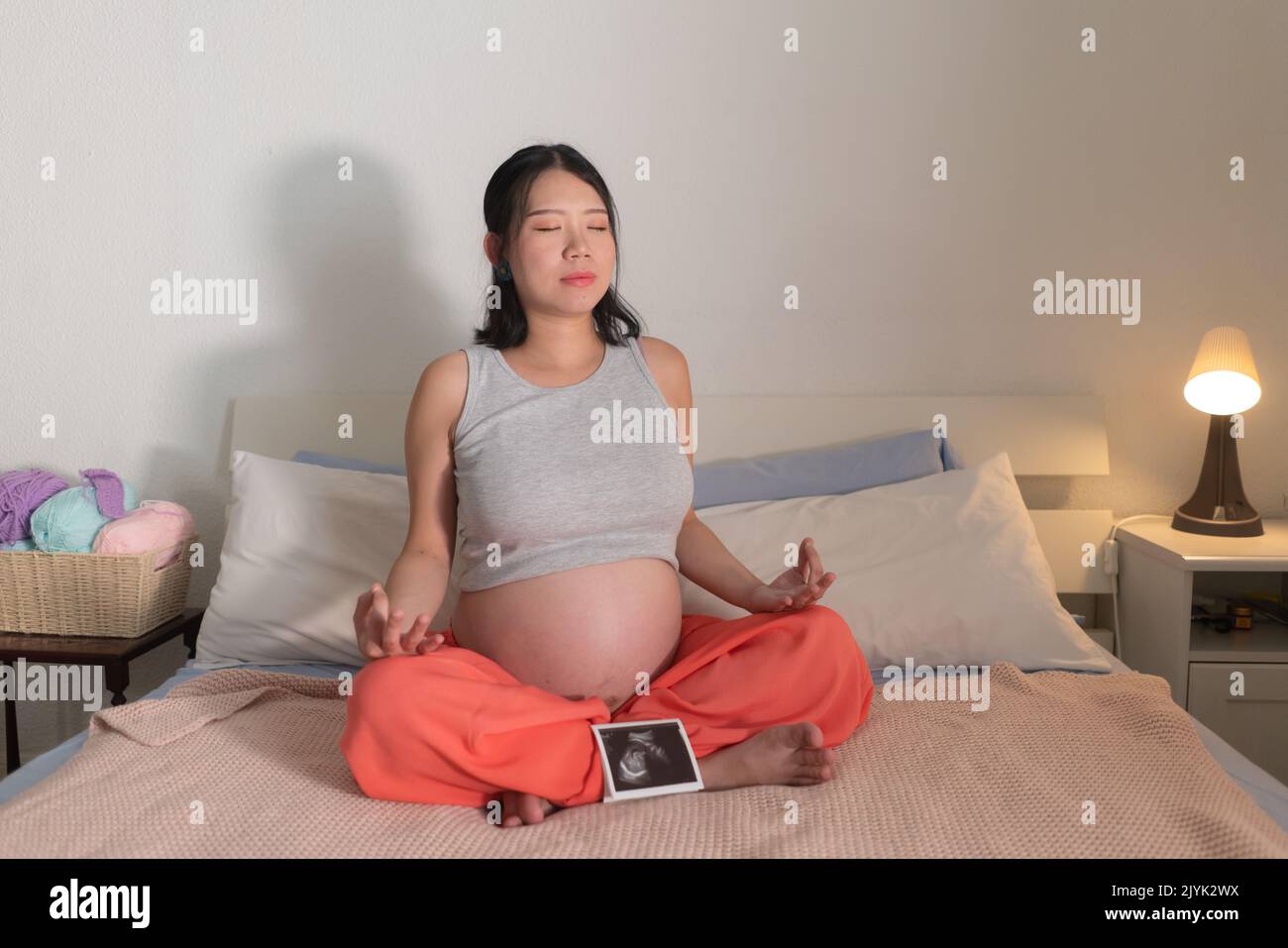 lifestyle home portrait of young happy and beautiful Asian Korean woman pregnant doing relaxing yoga exercise on bed in maternity and pregnancy concep Stock Photo