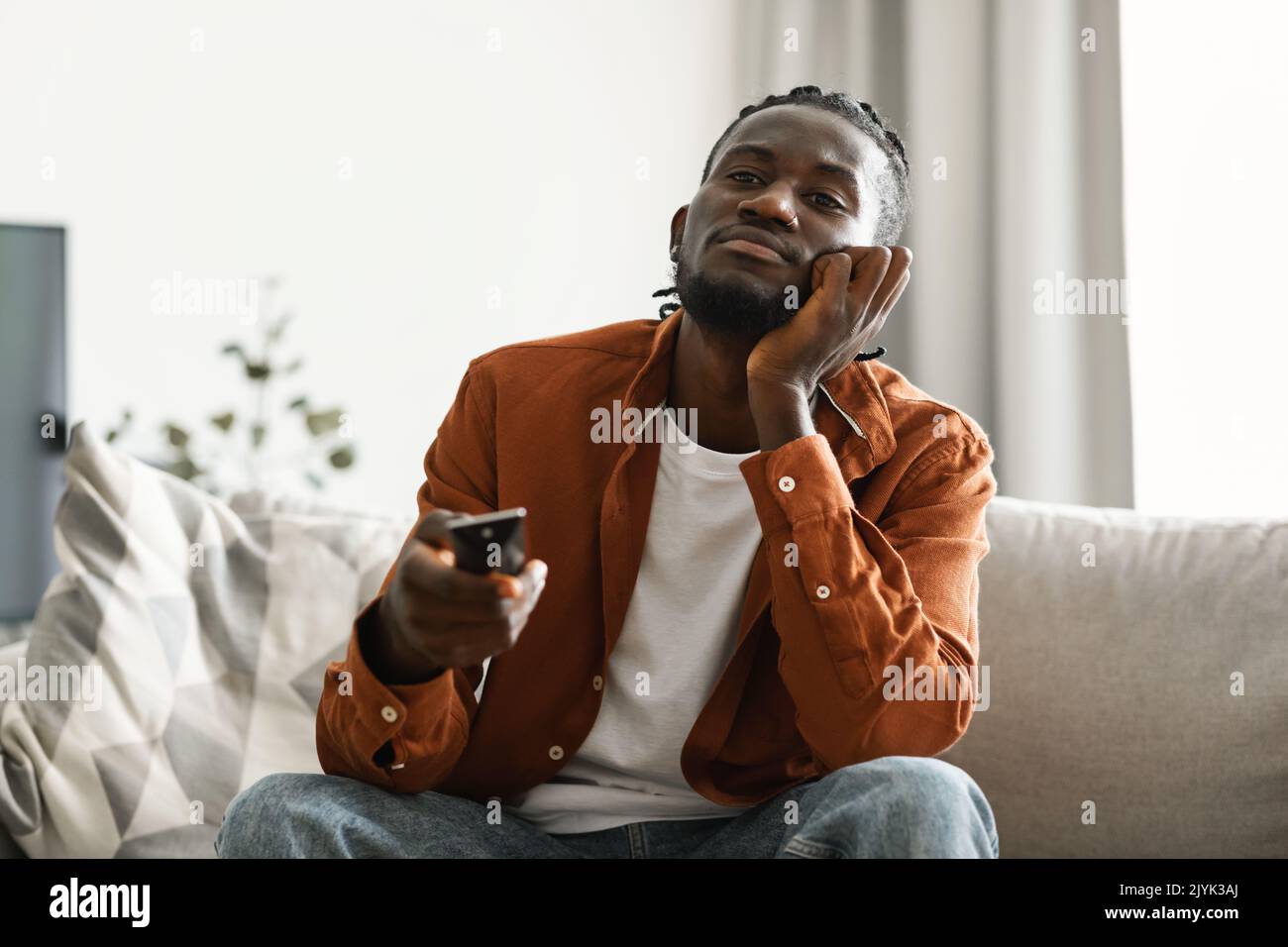 Boring television program. Bored black man watching TV and switching channels with remote control, sitting on couch Stock Photo