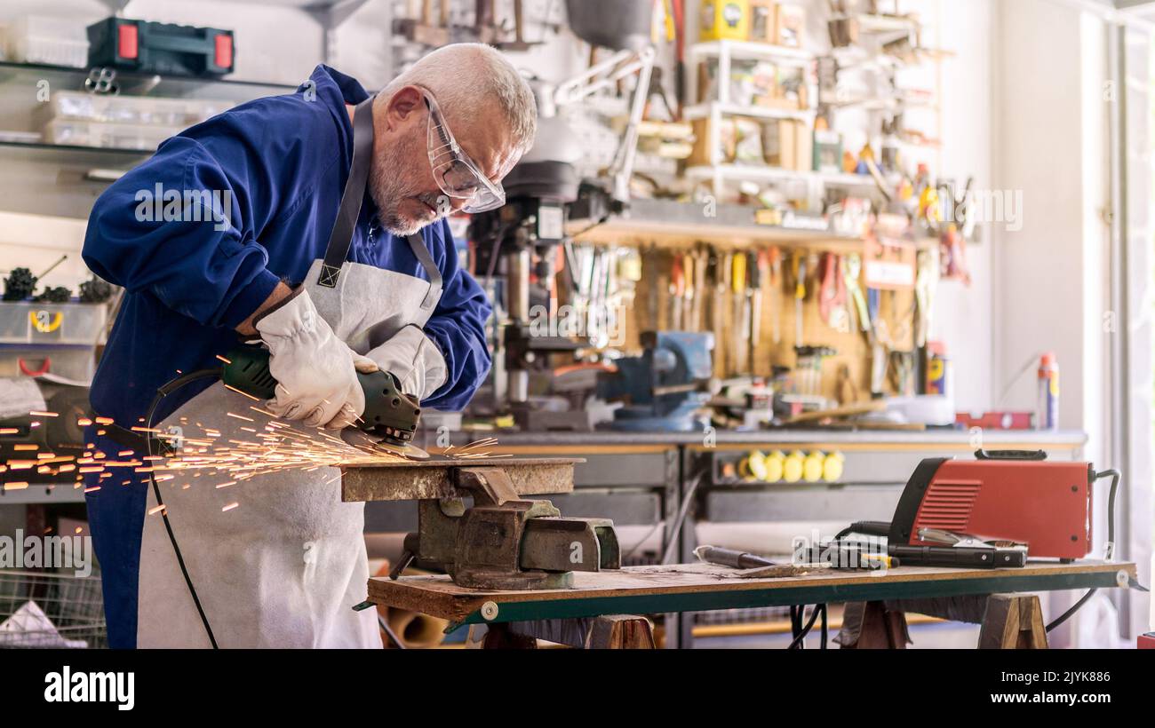 Worker in special clothes and goggles grinding metal with a circular saw in his workshop, bright sparks of metalworking fly in different directions. Stock Photo
