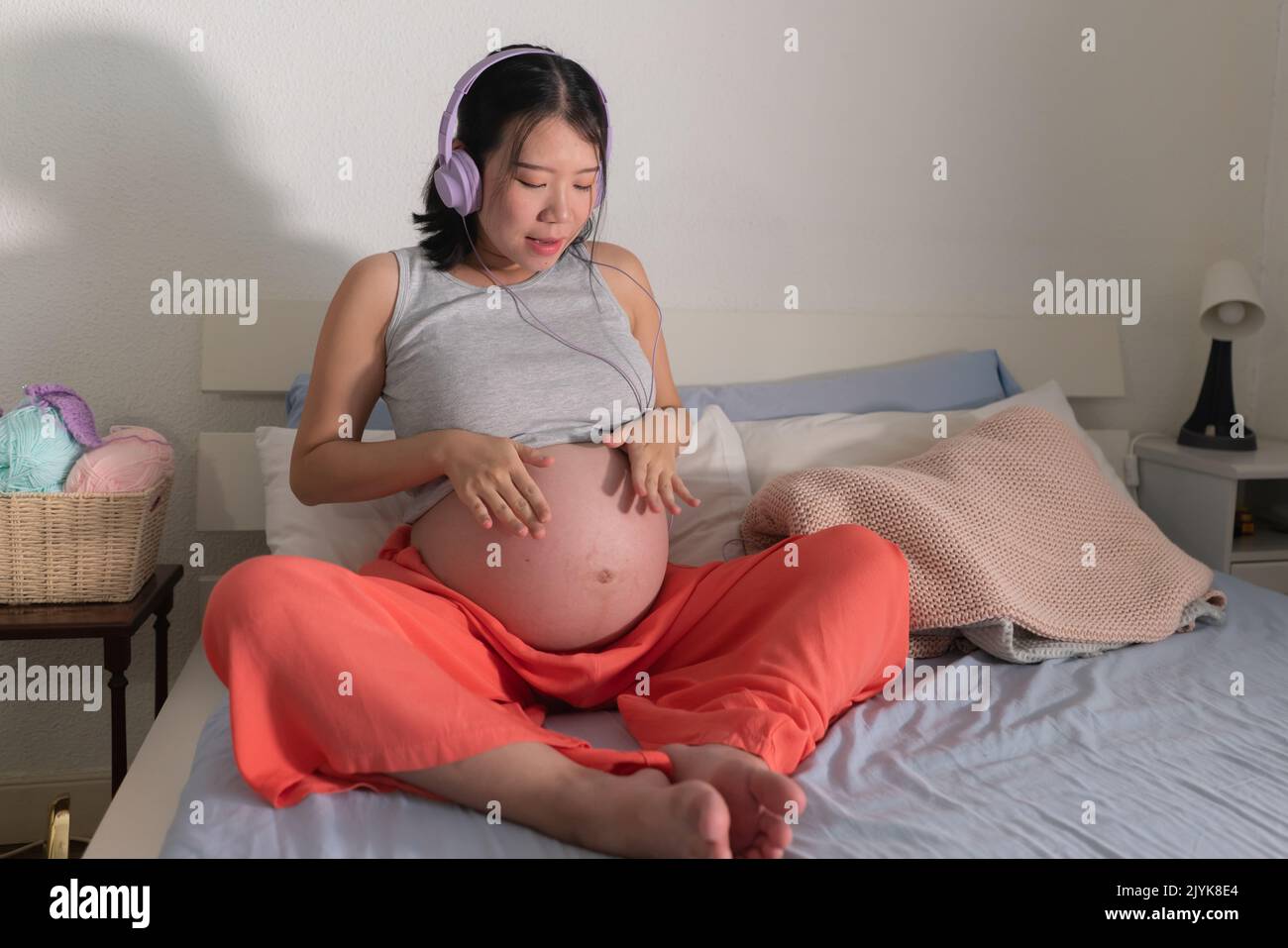 pregnancy lifestyle home portrait of young happy and beautiful Asian Chinese woman sitting pregnant on bed listening to music with headphones touching Stock Photo