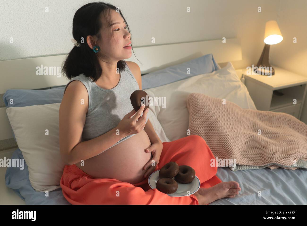 young happy and beautiful Asian Japanese woman pregnant sitting on bed holding dish full of chocolate donuts craving for sweet food in pregnancy conce Stock Photo