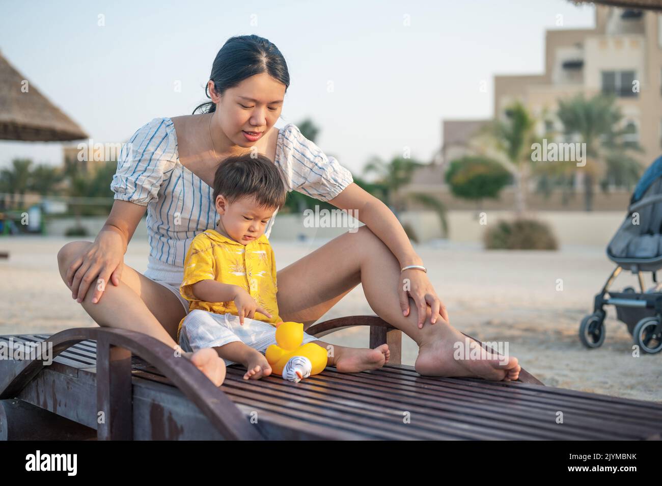 Baby boy on a beach holiday sitting on the sunbed with his mother and playing with toy duck. Asian woman and her one year old infant boy having fun on Stock Photo