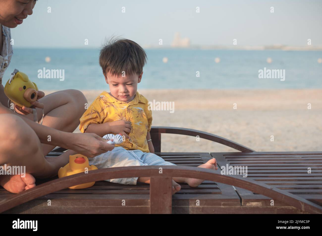 Baby boy on a beach holiday sitting on the sunbed with his mother and playing with toy duck. Asian woman and her one year old infant boy having fun on Stock Photo