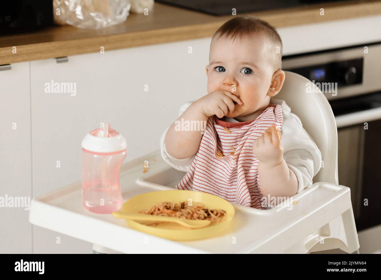 A young baby sitting in a high chair feeding herself spaghetti with a dirty face Stock Photo
