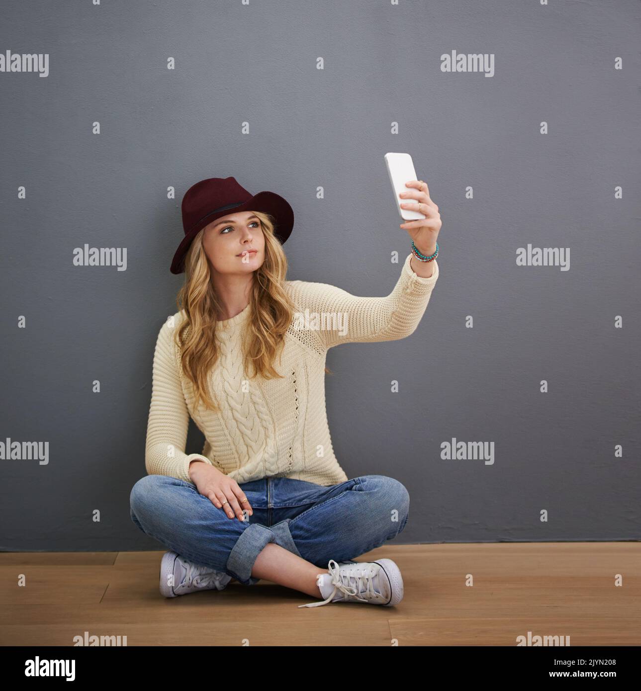 But first, lemme take a selfie. a young woman using her cellphone while sitting against a gray background. Stock Photo
