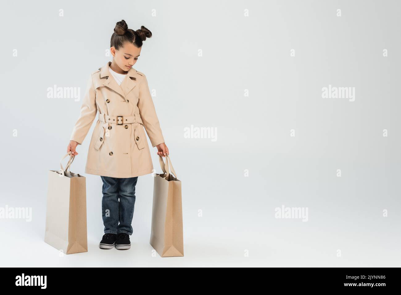 full length of stylish kid in trench coat and jeans holding shopping bags on grey, black friday concept,stock image Stock Photo