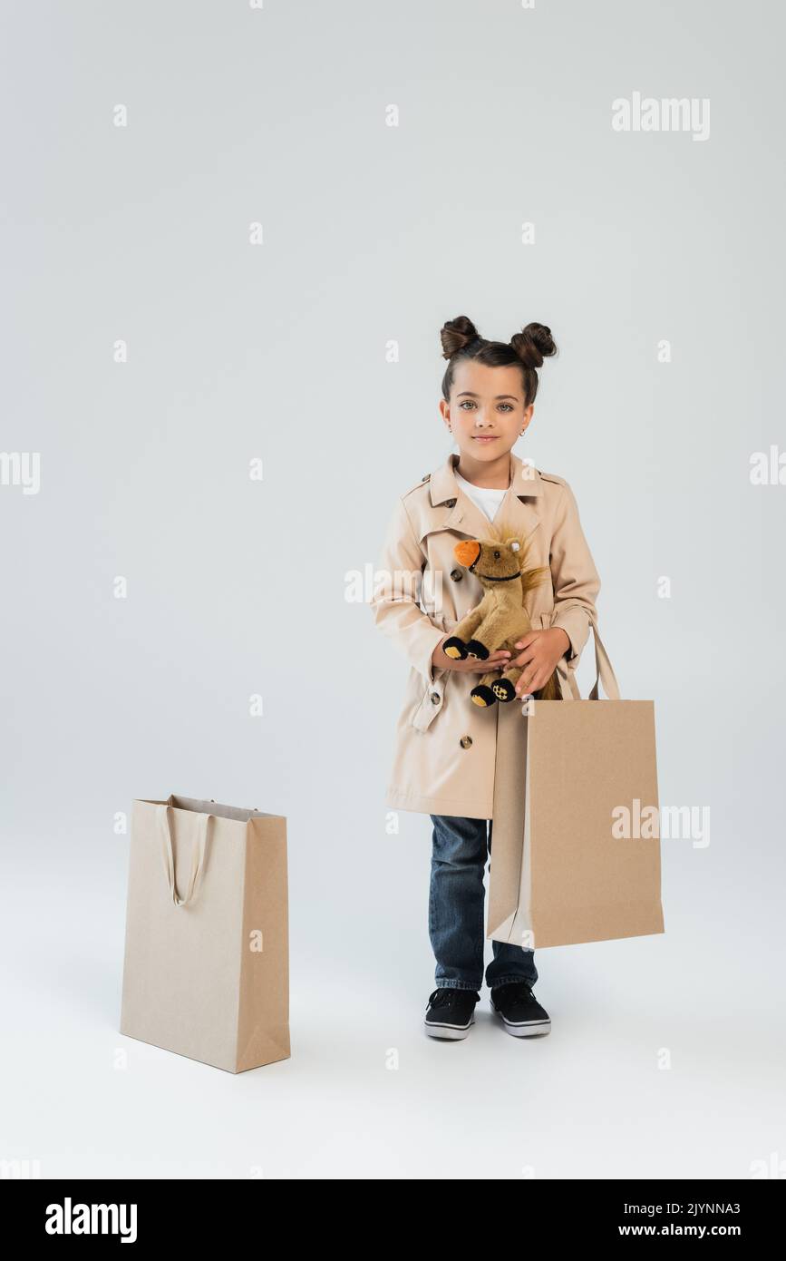 full length of cheerful kid in trench coat and jeans holding soft toy and shopping bag on grey,stock image Stock Photo