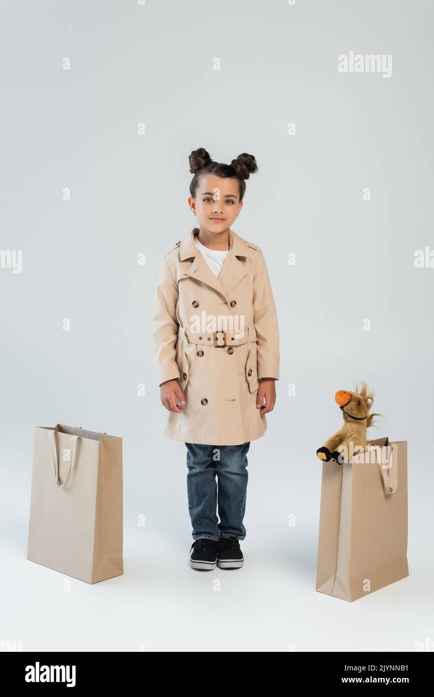 full length of cheerful kid in trench coat and jeans standing near shopping bags with soft toy on grey,stock image Stock Photo