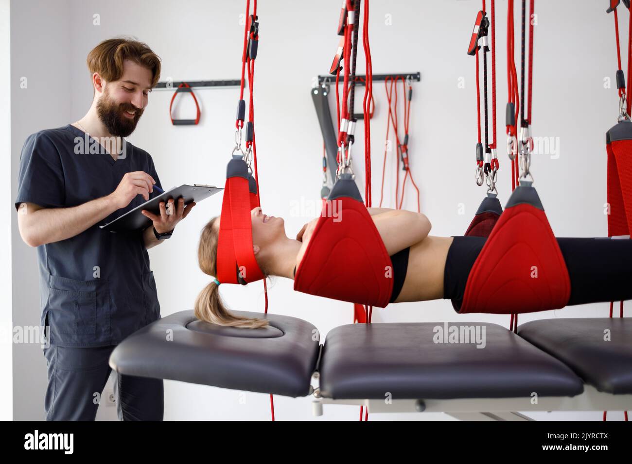 Physiotherapist doctor with clipboard watching female patient hanging on suspension system for neuromuscular activation in rehabilitation clinic Stock Photo