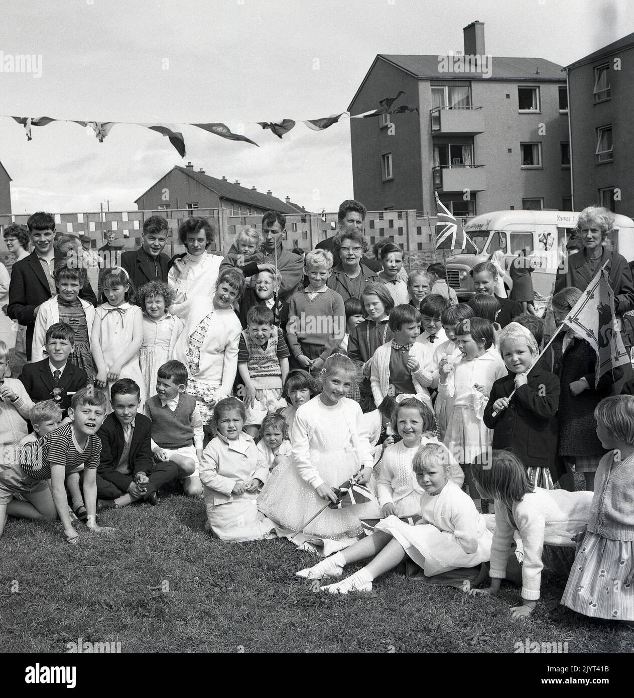 1965, historical, children and adults gathered for a group picture, some holding flags, including a union jack, after taking part in the North Queensferry gala day, Fife, Scotland, UK. A Mr 'whippy' ice-cream van of the era is seen in the background. Stock Photo