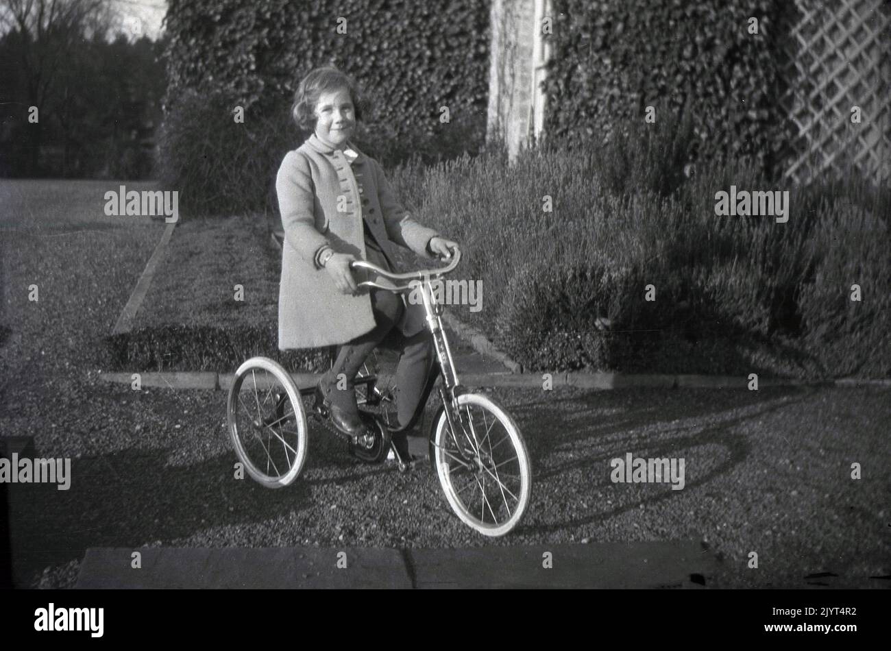 1934, historical, outside a country house on a gravel path, a young girl in a stylist coat and boots, sitting on a tricycle - possibly a Tri-ang model made by the British company, Lines Bros- England, UK. Children's tricycles with a pedal and chain and equal size wheels - like the one seen here - began to become popular in the 1910s and WW1 saw major improvements to chain-driven design, created by the demand for invalid carriages for the injured soldiers. Stock Photo