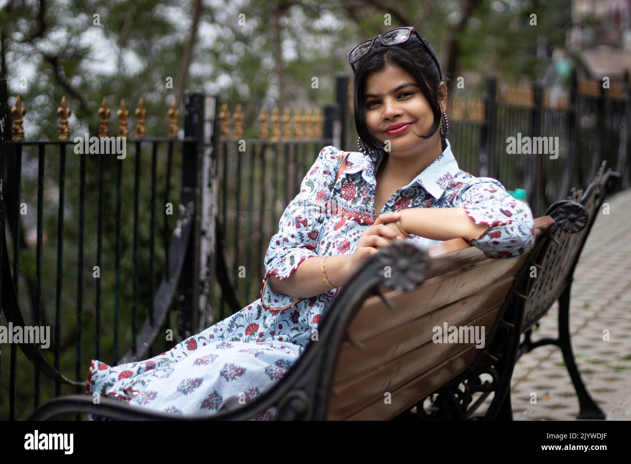A pretty Indian woman with sunglasses on head smiling and looking at camera while sitting on a park bench Stock Photo