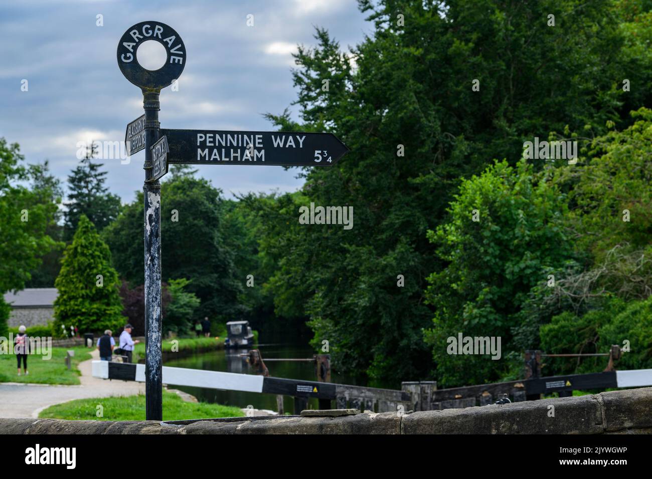 Waymark sign pointing to Pennine Way footpath route by canal lock & people walking - Leeds and Liverpool Canal, Gargrave, North Yorkshire, England UK. Stock Photo