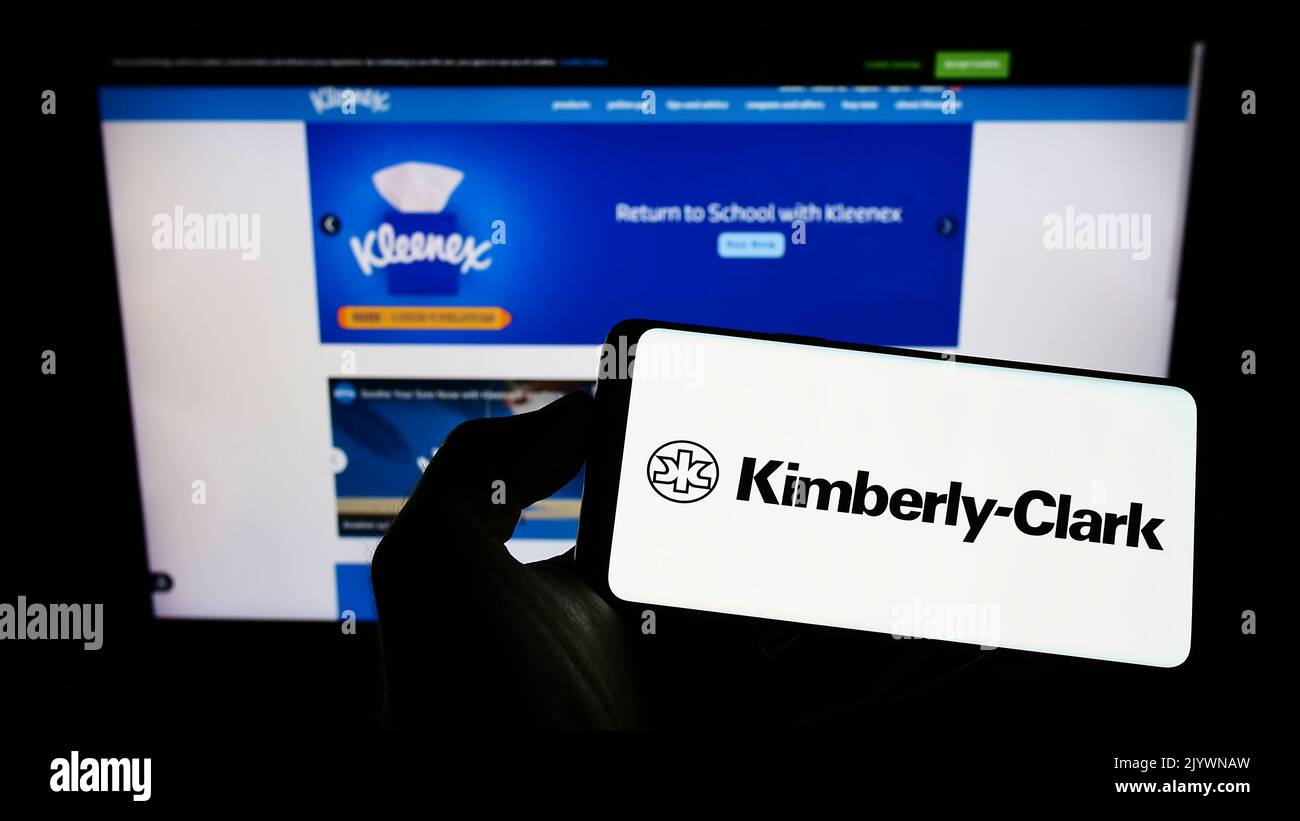 Person holding cellphone with logo of personal care company Kimberly-Clark Corporation on screen in front of webpage. Focus on phone display. Stock Photo