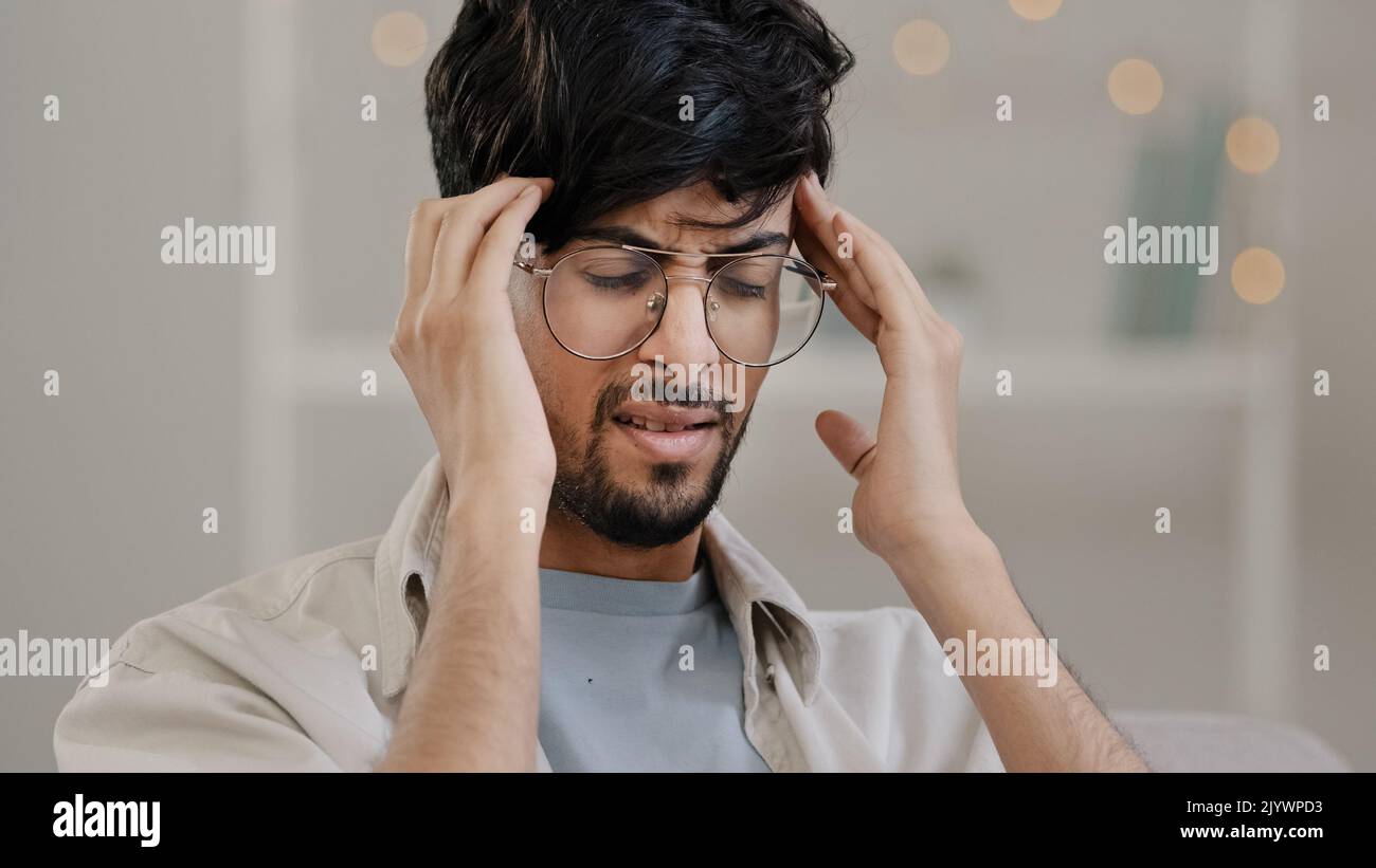 Close-up arabic man bearded guy with glasses feels stress overwork cranial pressure massages weight on head suffering from headache migraine sighing Stock Photo