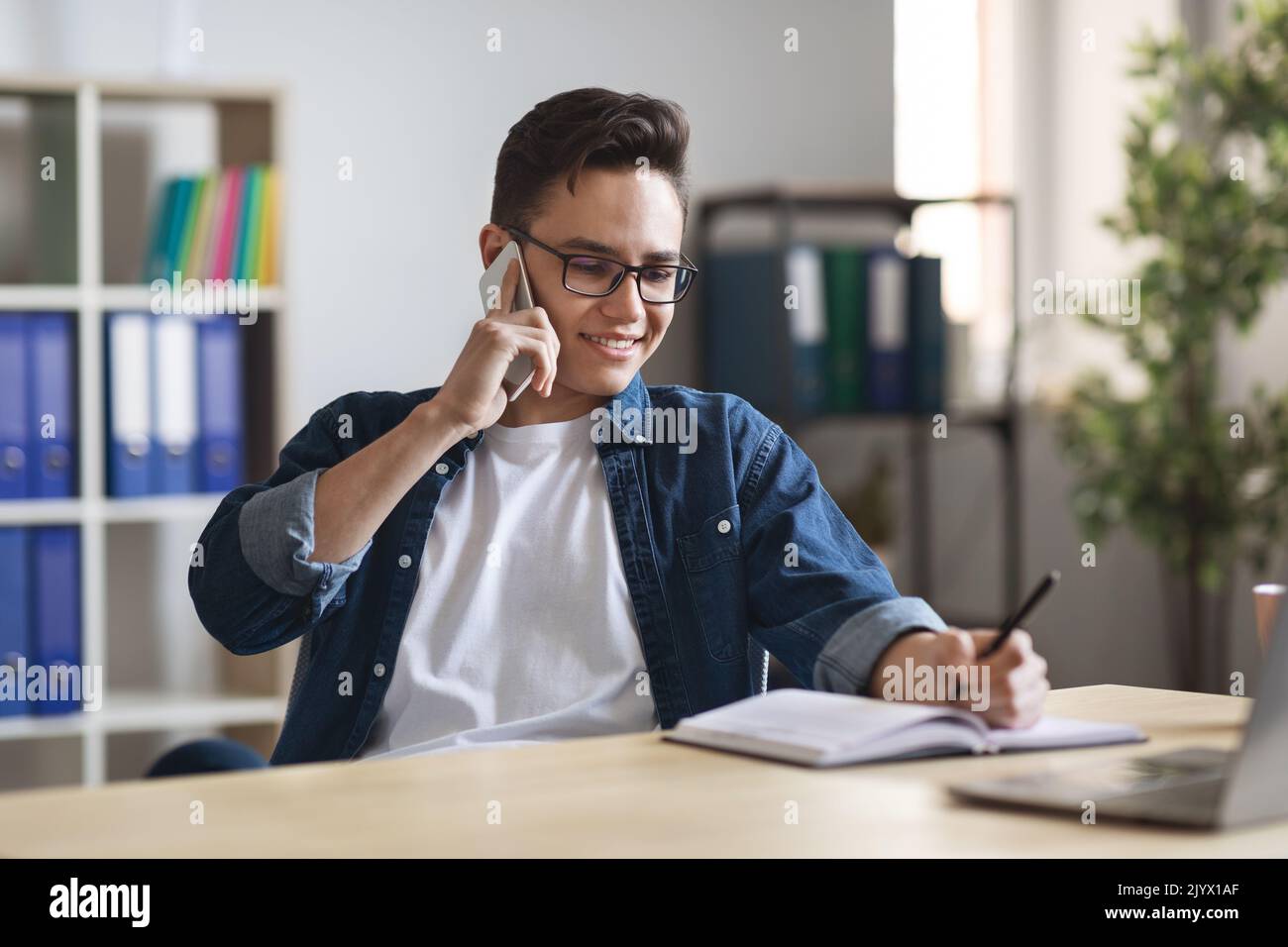 Millennial man writing notes while making phone call at workplace in office Stock Photo