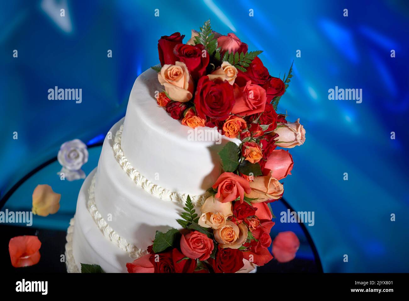 Wedding cake decorated with real roses. Cascading red and pink roses on an elegant three tier, white frosted cake.  Background is a dreamy deep blue Stock Photo