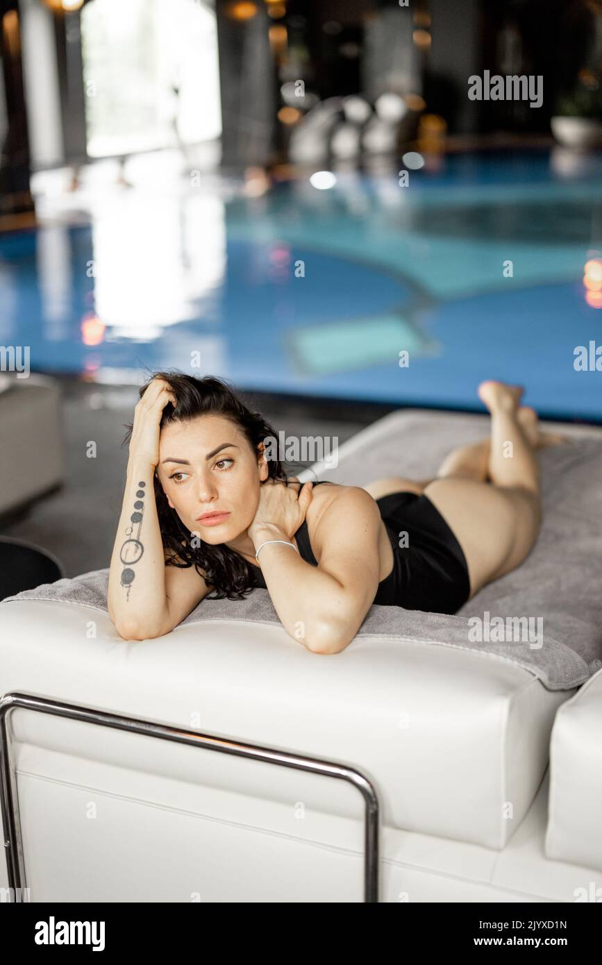 Portrait of adult woman in black swimsuit lying relaxed on lounge bed by the swimming pool. Woman spends holidays at spa resort with an indoor pool Stock Photo