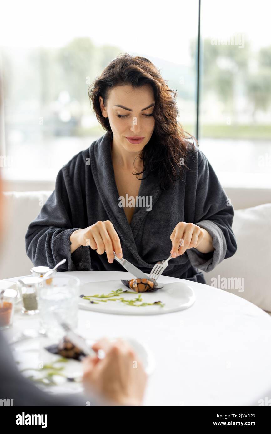 Couple eating beautiful haute cuisine dishes sitting by the table in restaurant at spa resort. Woman wearing bathrobe Stock Photo