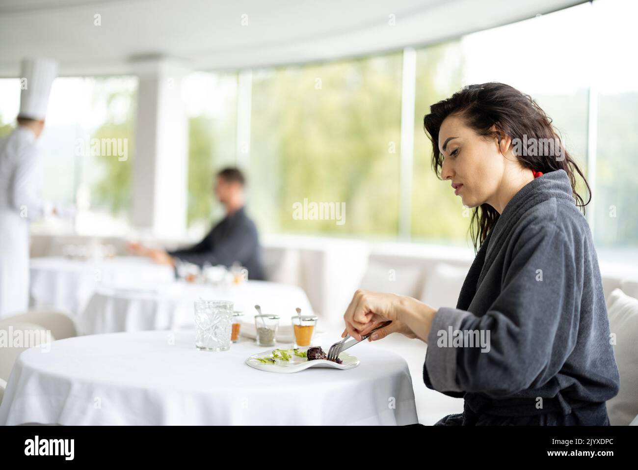 Beautiful woman in bathrobe enjoys delicious meal, sitting in white restaurant of haught cuisine during vacations at luxury spa resort Stock Photo