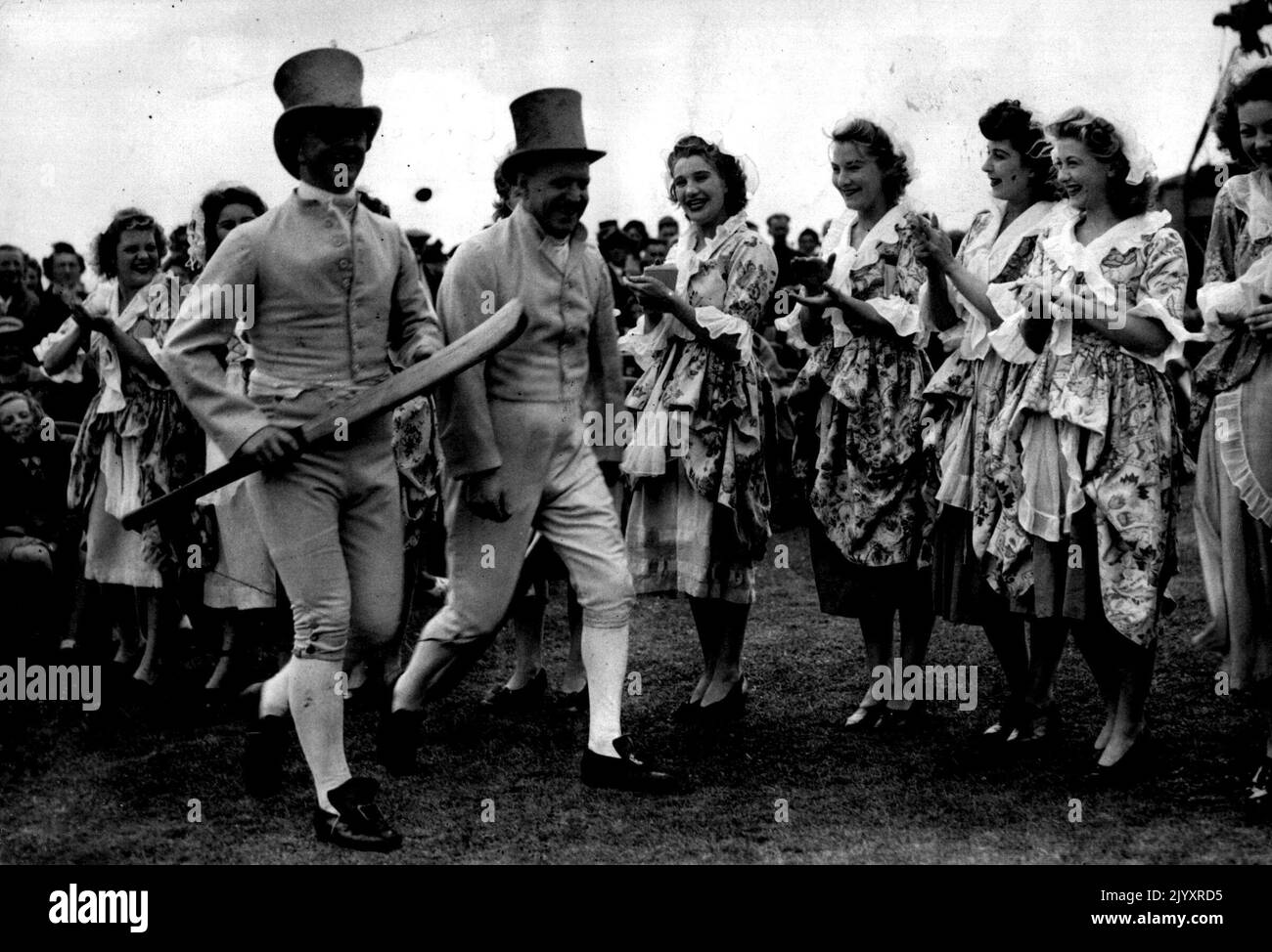 Cricket Puts The Clock Back -- The Regency Club's top-hatted and quaintly-attired opening batsmen get a clap from Regency ladies as they go out to bat. Their curved bats are in the style of a century ago. A cricket match in the style of a hundred years ago was played at Brighton to-day (Wednesday). Wickets of two stumps, bats and balls of the period, and top-hatted players were features of a challenge match between the Regency Club and the Hambledon Club, birth-place of cricket. Of the two scorers, one cut notches on a stick in the good, old-fashioned way, the other using modern scoring book i Stock Photo