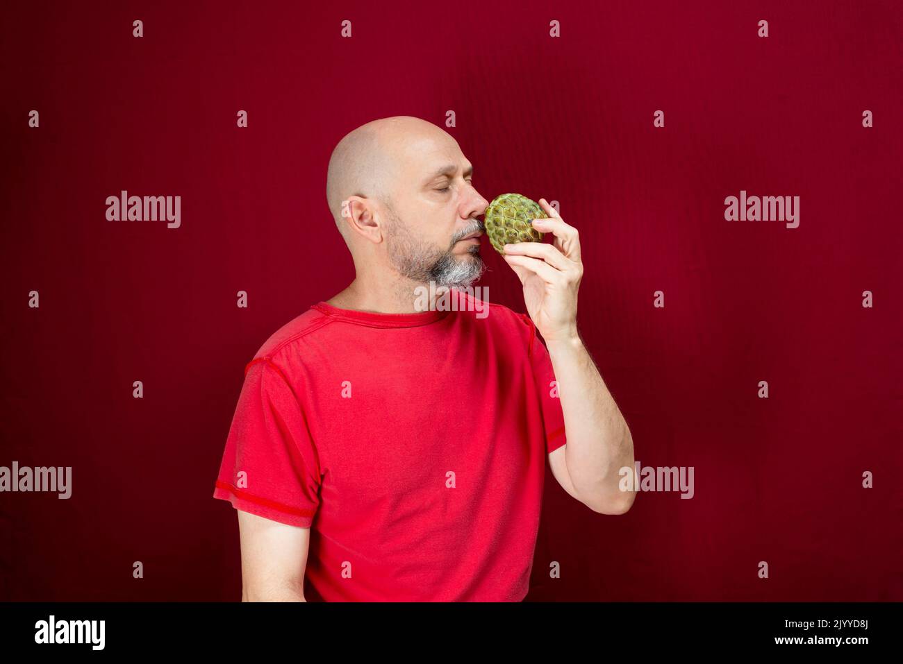 Young handsome man with beard, bald head and red shirt standing holding pinecone fruit against red background. Positive and healthy person. Stock Photo